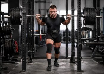 Heavy squat setup by competitive powerlifter with loaded barbell, lifting belt, and knee sleeves inside a gym power rack before unracking the weight.