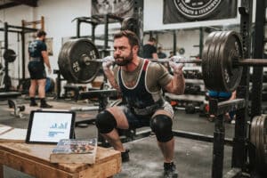 A focused powerlifter performing a heavy barbell back squat in a gritty gym, with a tablet displaying workout research data and training notebooks resting on a wooden bench nearby, bridging scientific data with practical strength training.